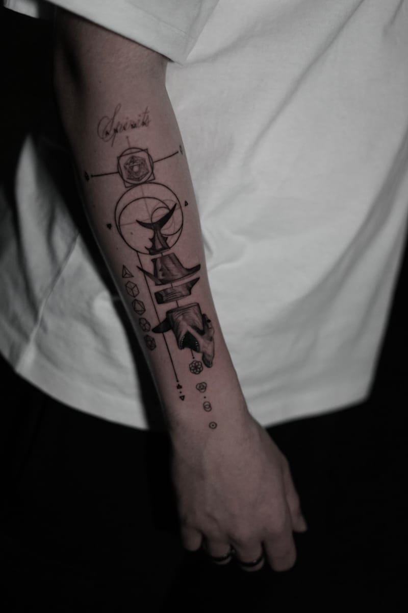 Antebrazo con un tatuaje vertical en línea fina: la palabra Spiritus en caligrafía sobre las letras ANIMA, un icosaedro dentro de un cuadrado, una cola de ballena sobre un círculo y una cabeza de tiburón, sobre camiseta blanca.