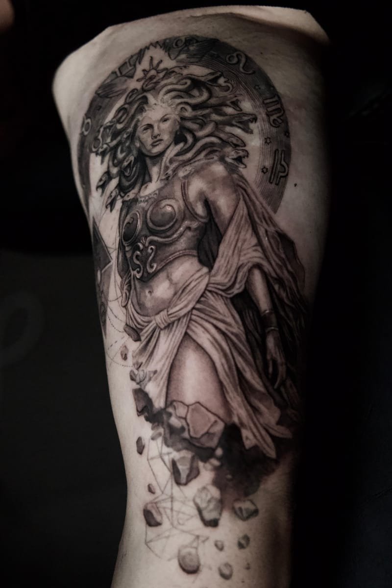 Tatuaje en negro y gris de una figura femenina con serpientes en el cabello, vestida con túnica y peto, de pie ante un disco con símbolos del zodiaco; bajo ella se desprenden fragmentos de piedra.