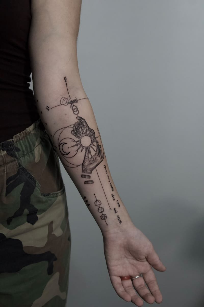 Antebrazo completo sobre fondo gris, del codo a la muñeca: dos manos sostienen un sol radiante junto a una luna creciente entre círculos, con símbolos alquímicos arriba y, hacia la muñeca, tres poliedros y la frase «Deus non ludit alea cum uni verso.»