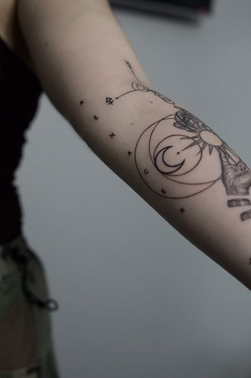Detalle de la cara interna del codo: las letras de la palabra «renacer» rodean la luna creciente y sus círculos; a la derecha asoman el sol y una de las manos.