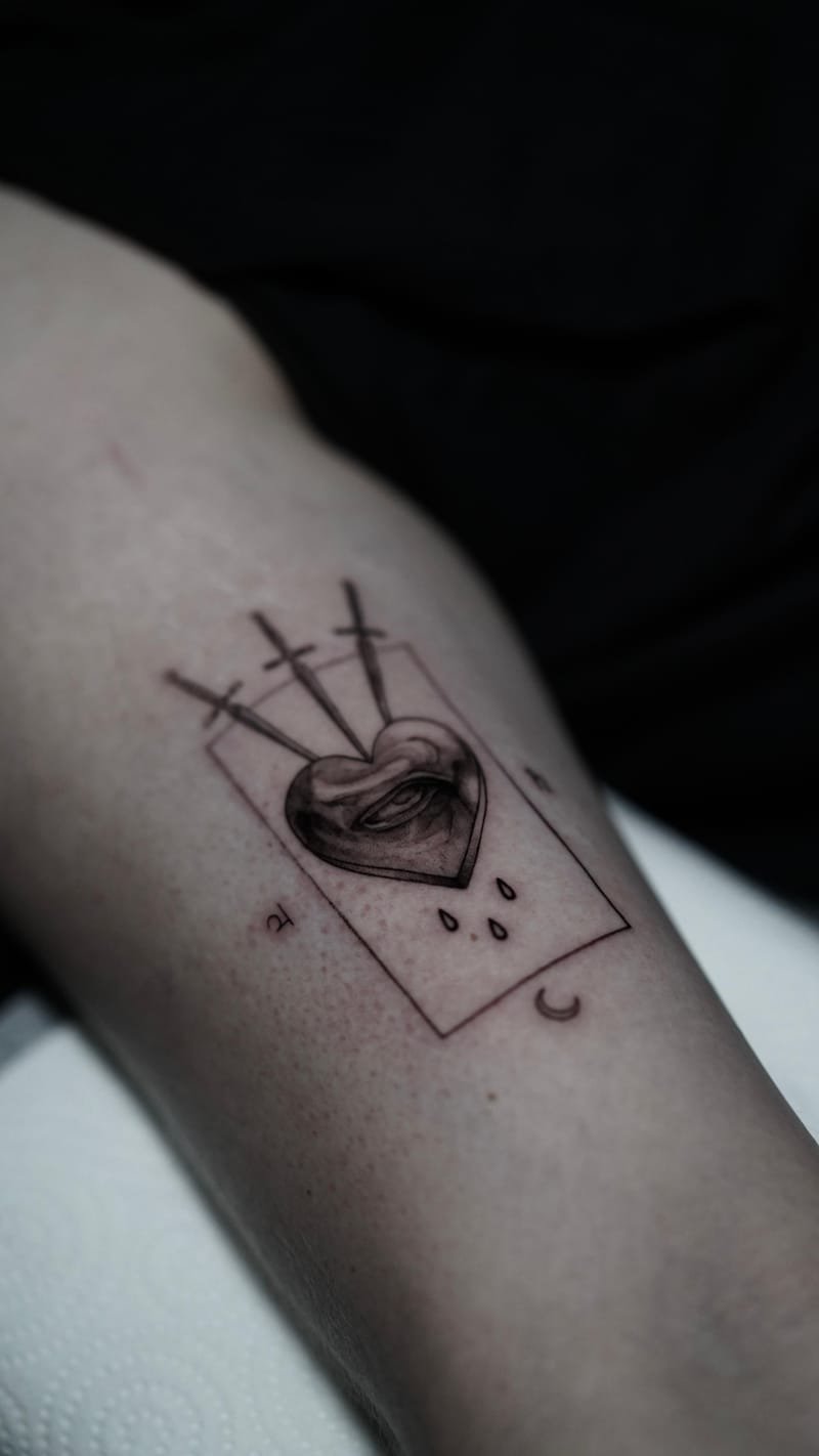 Tatuaje en la cara interna del brazo de la carta Tres de espadas: un corazón sombreado en gris con un ojo entreabierto en el centro, atravesado por tres espadas finas, sobre el marco rectangular de una carta; debajo, tres gotas y una pequeña luna creciente.